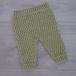 Könnte beinhalten: Eine grüne, gehäkelte Babyhose. Die Hose hat ein strukturiertes Muster und ist aus weichem Garn gefertigt.