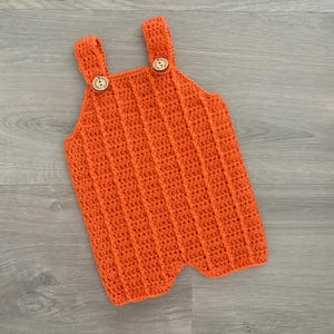 Könnte beinhalten: Orangefarbene gehäkelte Baby-Latzhosen mit Holzknöpfen.