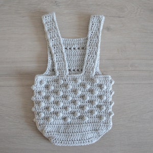 May include: A gray crocheted baby romper with a textured, bobble stitch pattern. The romper has straps that go over the shoulders.