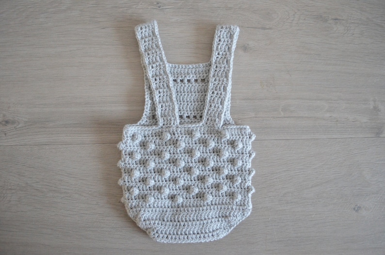 May include: A gray crocheted baby romper with a textured, bobble stitch pattern. The romper has straps that go over the shoulders.