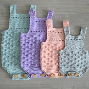 May include: Four crocheted baby onesies in different colors: light blue, purple, pink, and gray. Each onesie has a textured, bobble stitch pattern and wooden buttons.