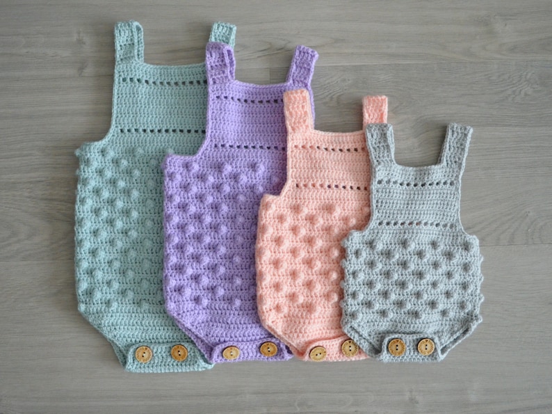 May include: Four crocheted baby onesies in different colors: light blue, purple, pink, and gray. Each onesie has a textured, bobble stitch pattern and wooden buttons.