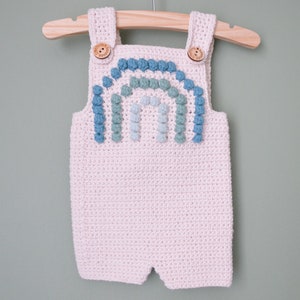 May include: A crocheted baby romper with a rainbow design made of blue and green pom-poms. The romper is white and has straps.