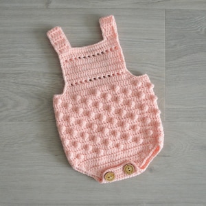 May include: A pink crocheted baby onesie with a textured, bobble stitch pattern. The onesie has a sleeveless design and features two wooden buttons at the bottom.