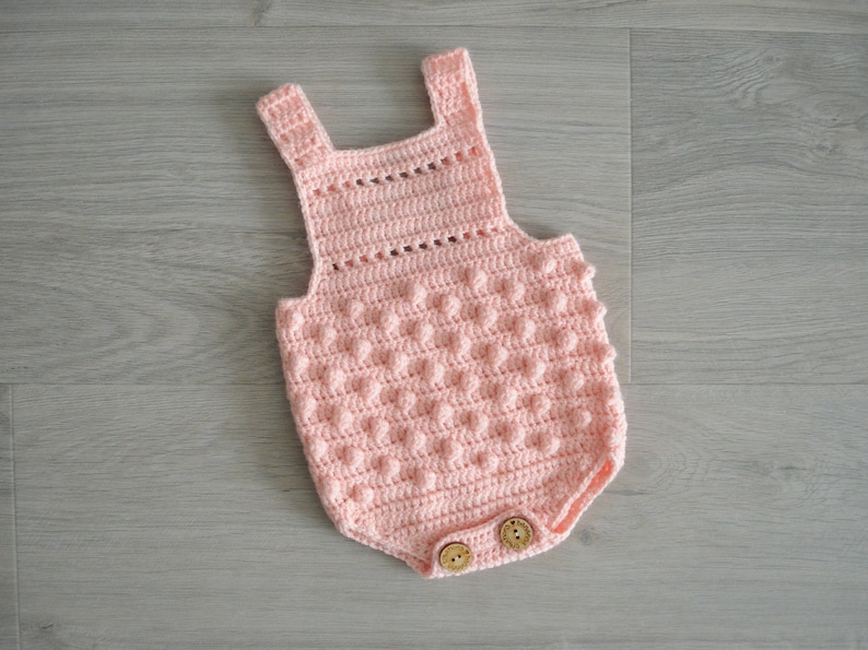 May include: A pink crocheted baby onesie with a textured, bobble stitch pattern. The onesie has a sleeveless design and features two wooden buttons at the bottom.