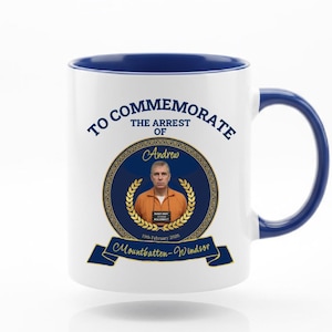 May include: White ceramic mug with a blue interior and handle. The mug features the text "TO COMMEMORATE THE ARREST OF Andrew" around a circular image of a man in an orange jumpsuit. Below the image is a blue banner with the text "Mountbatten-Windsor".