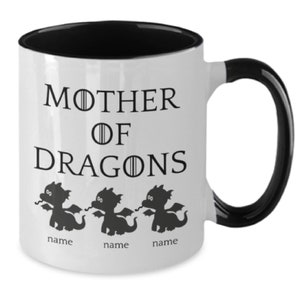 May include: White ceramic mug with a black rim and handle. The mug features a black silhouette of three dragons with the text "Mother of Dragons" and the word "name" beneath each dragon.