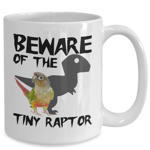 May include: White ceramic coffee mug with a black and grey illustration of a parrot and a dinosaur. The text "Beware of the Tiny Raptor" is printed on the mug.