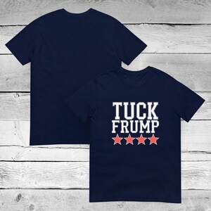 May include: Navy blue t-shirt with the text "TUCK FRUMP" in white block letters and four red stars. The shirt is displayed with the front and back visible, showcasing the solid navy color.