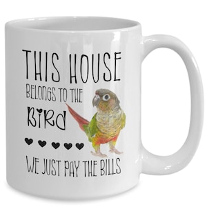 May include: White ceramic coffee mug with a black and white graphic of a green and red parakeet. The text on the mug reads "This house belongs to the bird. We just pay the bills."