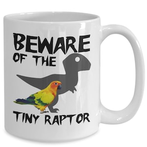 May include: White ceramic mug featuring the text "BEWARE OF THE TINY RAPTOR" in black. A colourful parrot is depicted below the text, with a grey dinosaur silhouette in the background. The mug has a large handle.