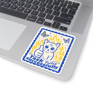 May include: A rectangular sticker with a blue border and the text "Task Failed Successfully." The sticker features a cartoon cat giving a thumbs up, with yellow flames and butterflies in the background.