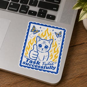 Puede incluir: Una pegatina rectangular con borde azul festoneado que muestra un gato blanco haciendo un gesto de pulgar hacia arriba frente a llamas amarillas. Dos mariposas azules y amarillas están sobre el gato. El texto dice Task Failed Successfully.