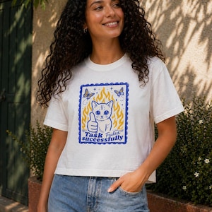 Puede incluir: Camiseta blanca con un diseño azul y amarillo de un gato con el pulgar hacia arriba, llamas y mariposas. El texto dice "Task Failed Successfully". La camiseta se combina con vaqueros azules.