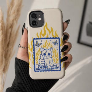 Puede incluir: Funda de teléfono crema con un gato de dibujos animados con el pulgar hacia arriba, rodeado de llamas amarillas y mariposas. El texto "Task Failed Successfully" está debajo.