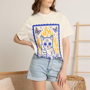 Puede incluir: Camiseta color crema con un gráfico azul y amarillo de un gato haciendo una señal de aprobación, rodeado de llamas y mariposas. El texto "Task Failed" está debajo del gato. La camiseta se combina con pantalones cortos vaqueros.
