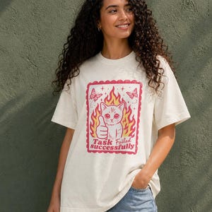 Puede incluir: Camiseta color crema con un gráfico de un gato haciendo una señal de pulgar hacia arriba, rodeado de llamas y mariposas. El texto "Task Failed Successfully" está debajo del gato. La camiseta es de corte holgado.