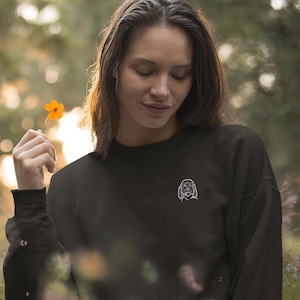 Könnte beinhalten: Eine Frau trägt einen schwarzen Pullover mit einem weißen gestickten Hundemuster und steht in einem Feld voller Wildblumen. Die Blumen sind in den Farben Rosa, Orange und Gelb.