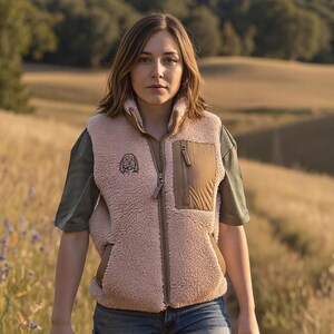 May include: A light pink fleece vest with a brown pocket and a small dog logo, worn over an olive green short-sleeved shirt. The vest has a full zipper. The person is wearing denim shorts and holding a red leash, walking on a path.