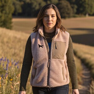 Può includere: Una donna indossa un gilet in pile rosa chiaro con una silhouette di cane nero, un maglione verde scuro e jeans blu scuro. Cammina su un sentiero, tenendo un guinzaglio rosso, con un campo sullo sfondo.