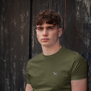 Könnte beinhalten: Ein Mann trägt ein grünes T-Shirt mit kurzen Ärmeln und einem kleinen weißen Hunde-Logo auf der linken Brust. Er trägt eine blaue Jeans und eine silberne und goldene Uhr.