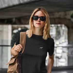 Pode incluir: Uma camiseta preta com uma pequena silhueta branca de um cão na frente. A pessoa que está usando a camiseta está usando óculos de sol e uma mochila marrom.