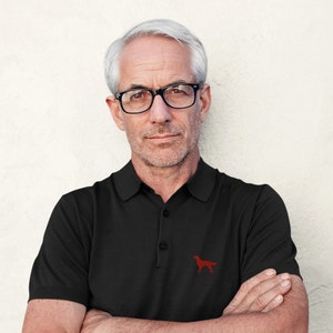 May include: A man with gray hair and a beard is wearing a black polo shirt with a red dog logo. He is wearing black-framed glasses and has a serious expression on his face.