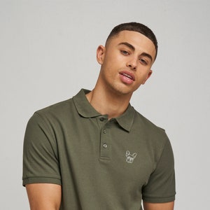 Puede incluir: Una camiseta polo de manga corta verde con una pequeña imagen bordada de un perro en el pecho izquierdo.