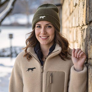 May include: A tan fleece jacket with a zip closure, a chest pocket, and two side pockets. The jacket features a black dog silhouette on the chest. The person is wearing a green beanie with a white dog silhouette.