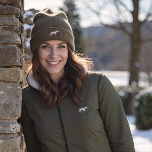 May include: An olive green jacket and beanie hat with a dog silhouette. The jacket has a zipper closure and a white fleece lining. The beanie hat is a knit style. The woman is smiling and leaning against a stone wall.