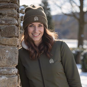 May include: An olive green jacket with a white fleece lining and a matching beanie. Both the jacket and beanie feature a small, embroidered emblem. The jacket has a full-length zipper and two pockets. The background shows a stone wall and a snowy landscape.