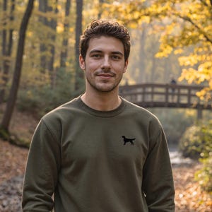 Può includere: Felpa verde oliva con una silhouette di cane nera sul petto. La felpa ha girocollo e polsini e orlo a coste. La persona indossa jeans blu scuro e tiene una corda marrone. Lo sfondo è una foresta con un ponte.