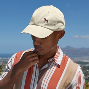 Könnte beinhalten: Beige Baseball Cap mit einer maroonfarbenen Hunde-Silhouette auf der Vorderseite. Die Kappe hat einen gebogenen Schirm und einen Stoffriemenverschluss. Der Hintergrund ist ein klarer blauer Himmel.