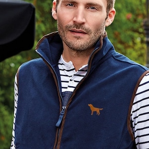 May include: A navy blue fleece vest with brown trim and a zipper closure. The vest has a brown dog silhouette embroidered on the front. The model is wearing a white and navy striped shirt underneath.