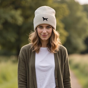 Puede incluir: Un gorro gris claro con una silueta de perro negro, usado con una rebeca verde, una camiseta blanca y vaqueros azules. El gorro es de punto acanalado. La mujer camina por un sendero.