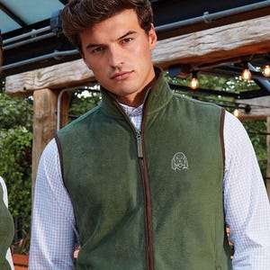 May include: Olive green fleece vest with a brown trim and a full zipper. The vest features a small embroidered design on the chest. The person is wearing a light blue and white checkered shirt underneath. The vest is a casual outerwear garment.