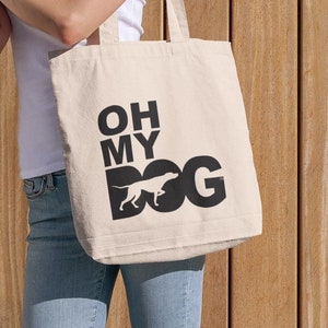 May include: A beige tote bag with the text "OH MY DOG" in black, featuring a dog silhouette. The bag is carried by a person wearing a white shirt and blue jeans. The background is a wooden wall.