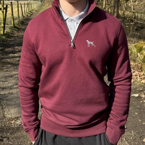 May include: A burgundy quarter-zip sweatshirt featuring a silver dog logo on the chest. The sweatshirt is worn over a grey polo shirt. The image shows a person wearing the sweatshirt.