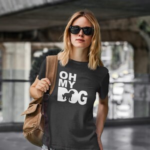 Può includere: T-shirt nera con la scritta bianca "OH MY DOG". La "O" di "DOG" è sostituita dalla sagoma di un cane. La persona indossa occhiali da sole e una borsa a tracolla marrone.