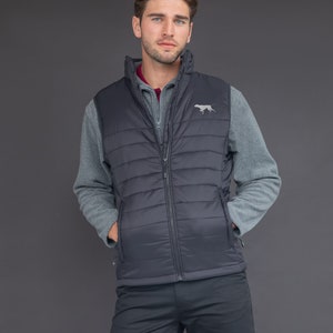 May include: A black quilted vest with a zipper closure and a small embroidered dog logo on the right chest. The vest is worn over a gray fleece pullover and black pants.