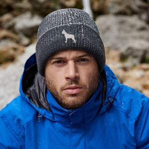 Op de afbeelding: Een man draagt een blauwe jas met capuchon en een grijze gebreide beanie met een wit honden silhouet logo.