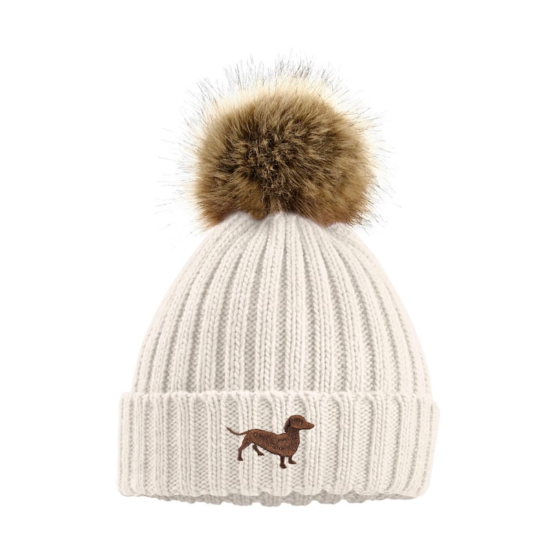 May include: White knitted beanie with a brown faux fur pom pom and an embroidered dachshund.
