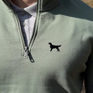 Può includere: Felpa verde salvia con zip a un quarto e una silhouette di cane nero sul petto. La felpa è indossata sopra una camicia grigia con colletto. La cerniera è parzialmente aperta. Lo stile è casual.