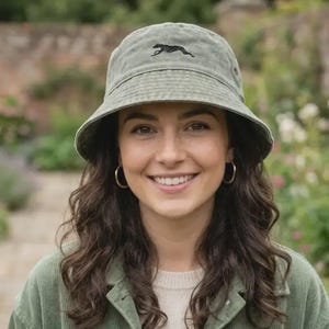 Könnte beinhalten: Ein salbeigrüner Fischerhut mit einem schwarzen, gestickten Geparden-Design. Der Hut ist aus einem weichen, strukturierten Stoff gefertigt und hat eine klassische, runde Form. Dieser Hut ist ein stilvolles Accessoire für Outdoor-Aktivitäten und Freizeitkleidung.