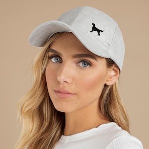 Può includere: Cappellino da baseball grigio chiaro con una silhouette di cane nero sul davanti. Il cappellino ha una visiera curva ed è realizzato in un materiale morbido e strutturato. Un accessorio alla moda.
