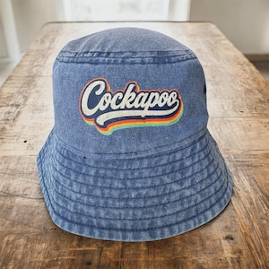 Puede incluir: Un sombrero de pescador de mezclilla azul con un gráfico de letras blancas desgastadas que dice Cockapoo, subrayado por una franja de arcoíris multicolor en rojo, naranja, amarillo, verde y azul. El sombrero tiene un ala cosida y una copa redondeada.