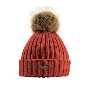 May include: A red knitted beanie with a brown faux fur pom pom on top. The beanie has a small embroidered dachshund on the front.