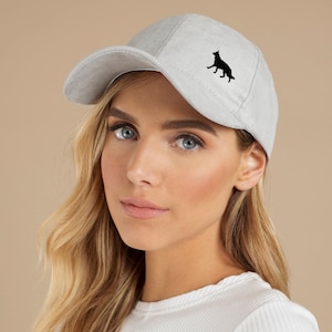 Op de afbeelding: Lichtgrijze baseballcap met een zwart silhouet van een hond aan de zijkant. De cap heeft een gebogen klep en een klassiek zes-panelen ontwerp. De hoed is gemaakt van een zacht, getextureerd materiaal.