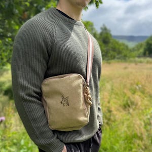 May include: A tan canvas crossbody bag with a black embroidered dog design. The bag has a striped strap and a rope detail on the side. The person is wearing a green sweater.