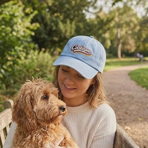 Può includere: Un berretto da baseball in denim azzurro con la scritta Cockapoo ricamata in corsivo, delineata da contorni sovrapposti marroni, arancioni e gialli. Il berretto presenta una visiera curva e una corona strutturata con un bottone centrale.
